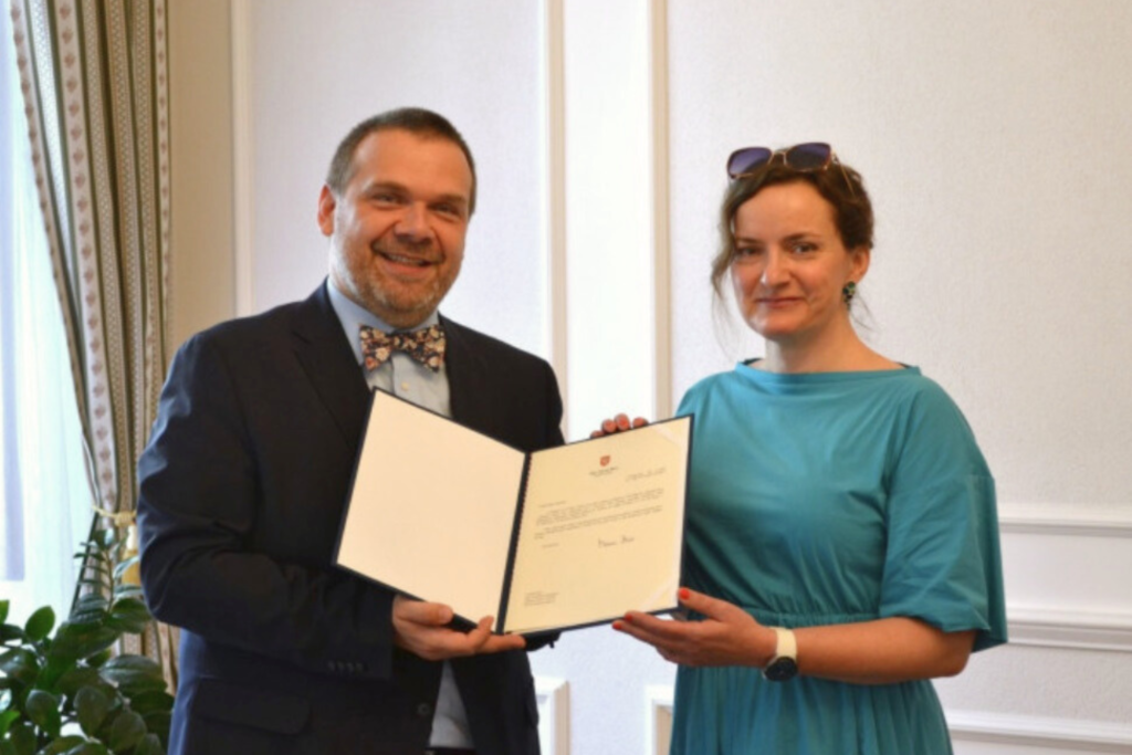 Žena v modrých šatech přijímá od muže v obleku pověření v deskách.