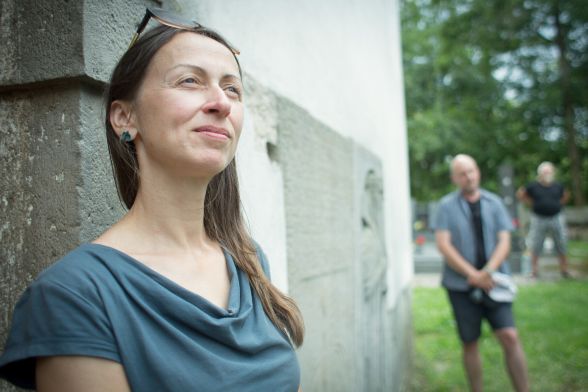 Žena stojící opřená o zeď. Za ní stojící muž před stromy.