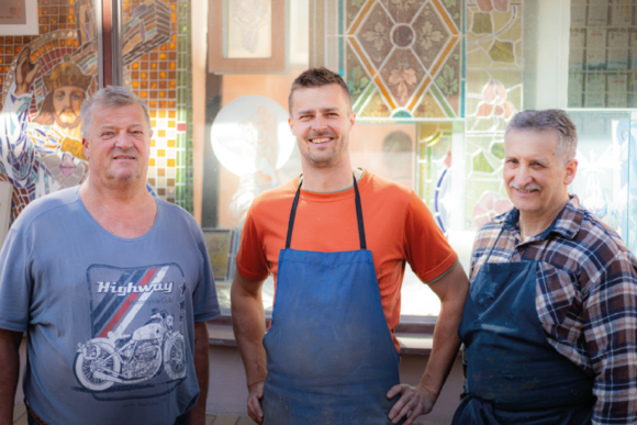 Tři muži stojí před skleněnými vitrážemi.