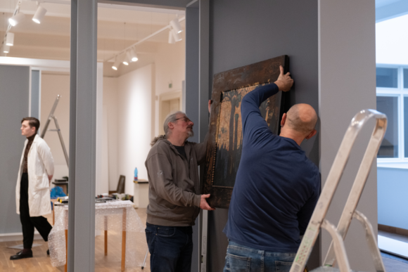 Dva muži věšící obraz. Další muž za nimi v bílém plášti si prohlíží obraz, který není vidět.